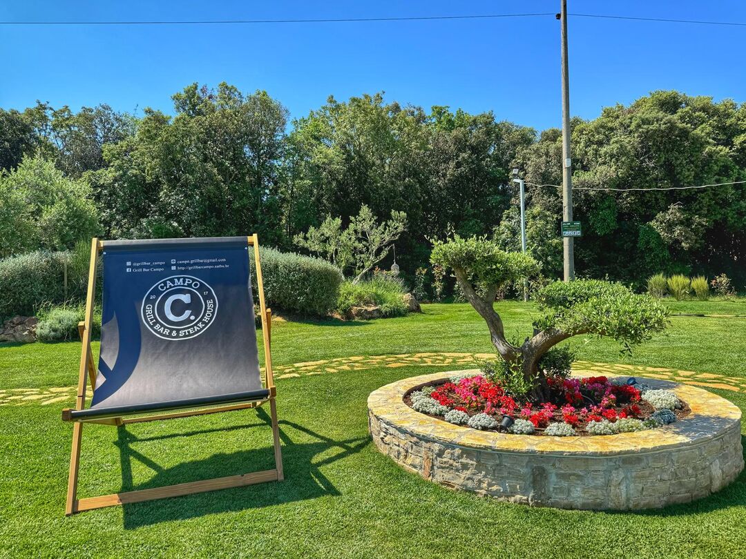Ležaljka s logotipom restorana Campo pokraj masline u kamenoj gredici s cvijećem