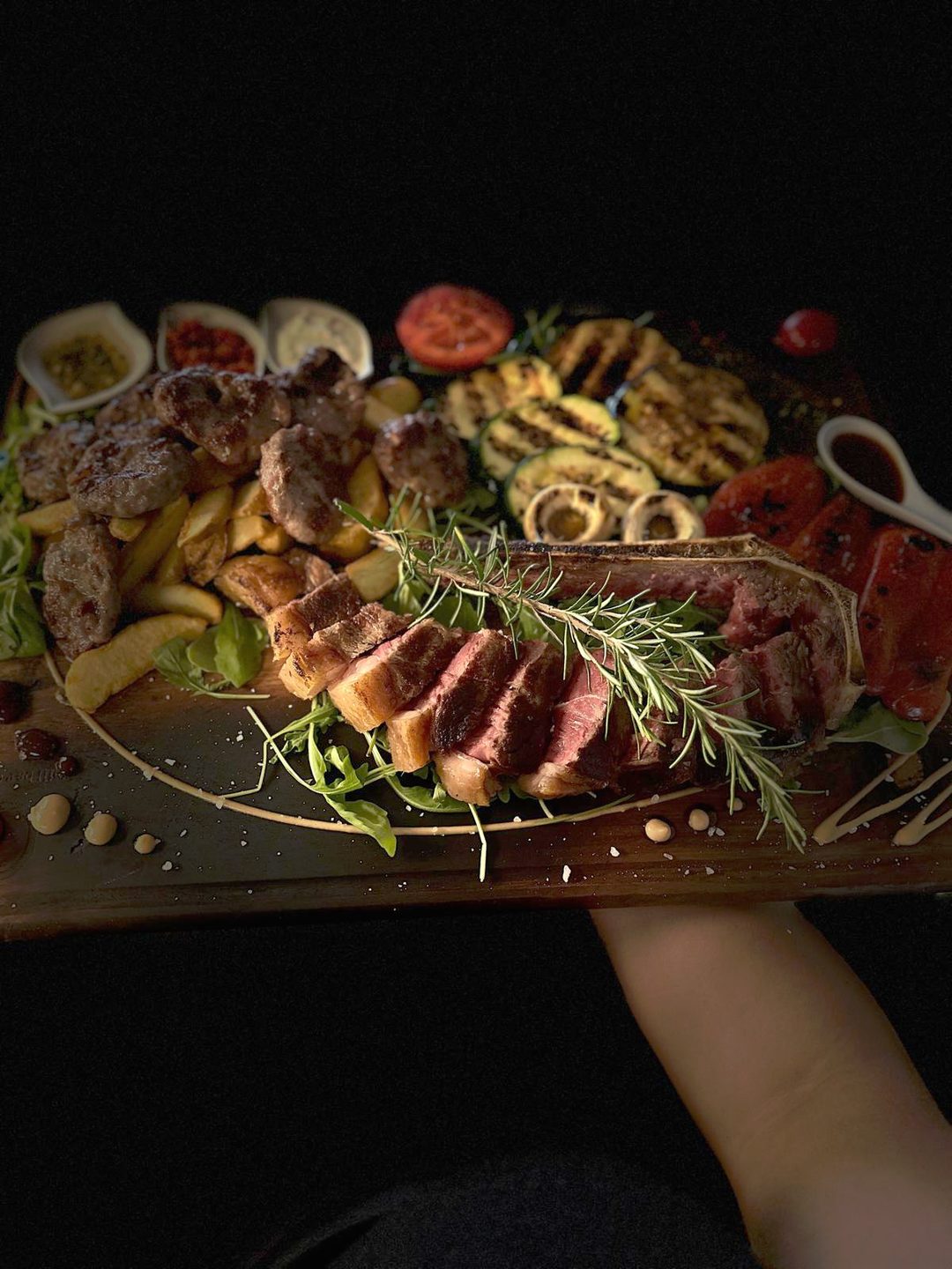Ruka nosi dasku s narezanim odreskom s kosti, ružmarinom, mesom i povrćem s roštilja